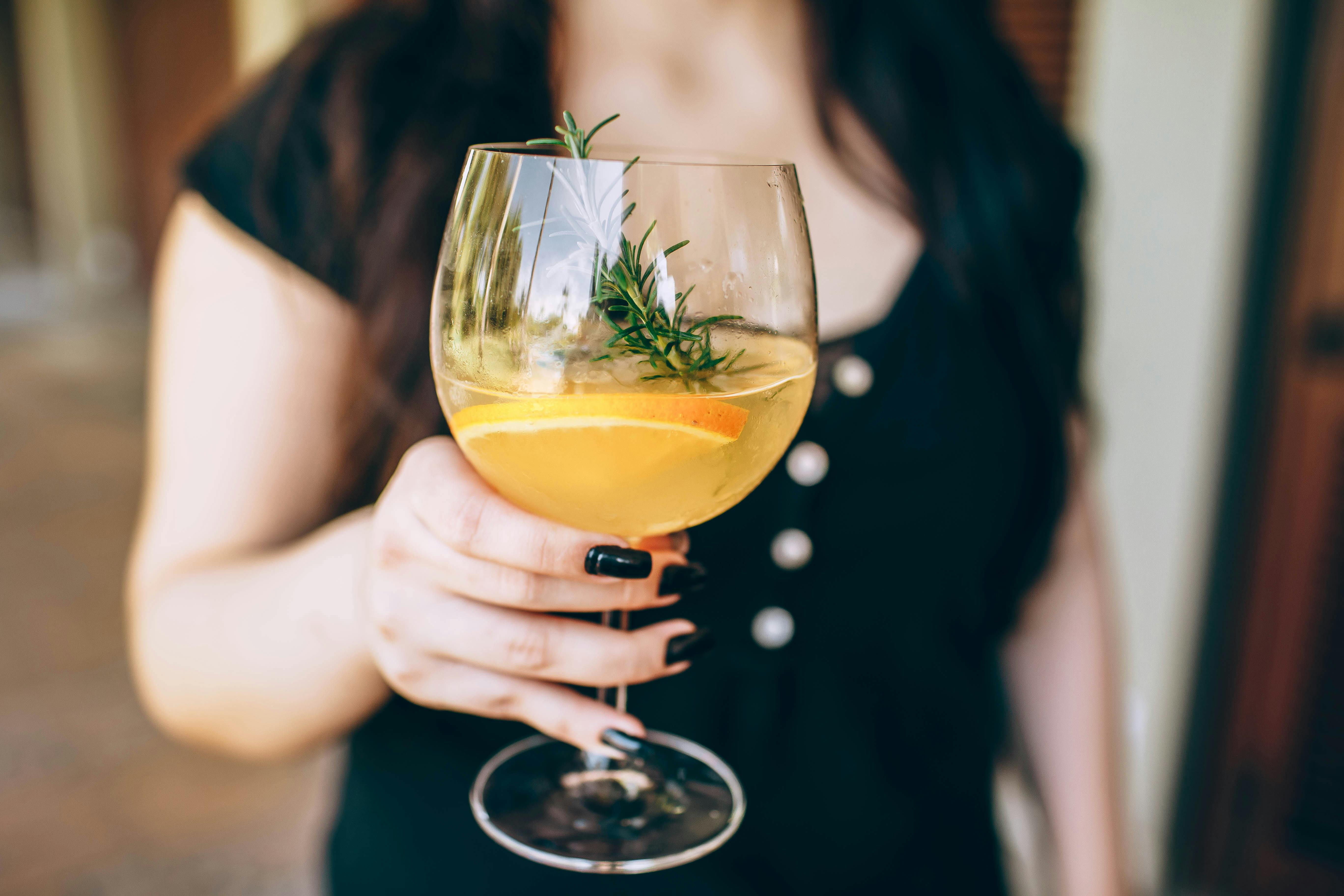 Close-up of a citrus cocktail in a wine glass held by a woman, garnished with rosemary.